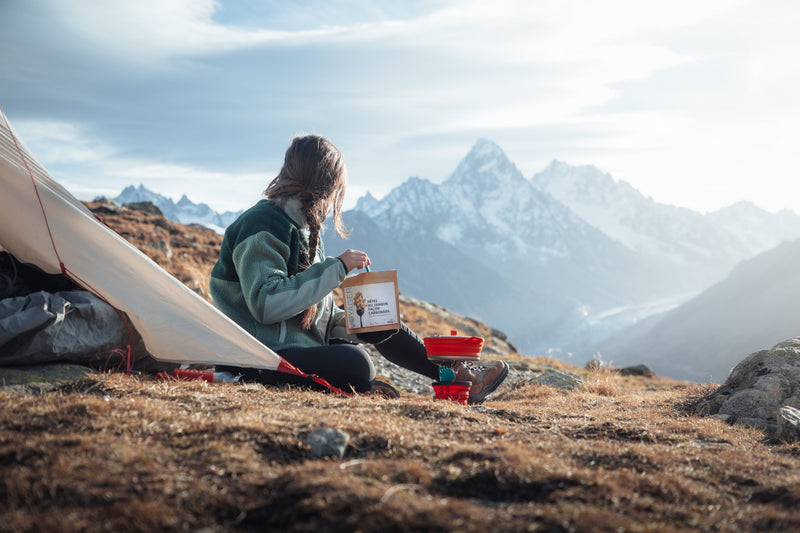 Randonneuse préparant un repas lyophilisé en bivouac face aux montagnes lors d’un trek outdoor